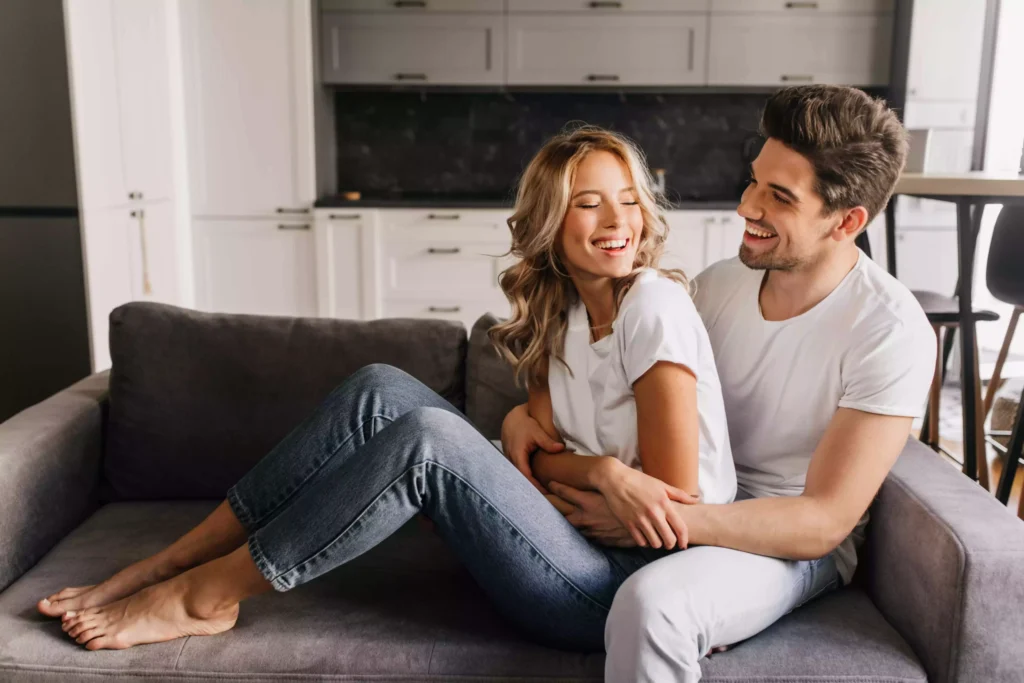 joyful day together in cozy and warm apartments happy attractive guy with beautiful girl looking at each other laughing and hugging on sofa 2 scaled 1